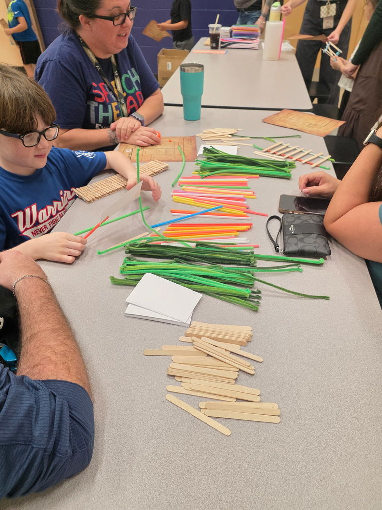 Two elementary students sitting at a cafeteria table, focused on building a structure using green pipe cleaners and wooden popsicle sticks during Storybook STEAM Night.
