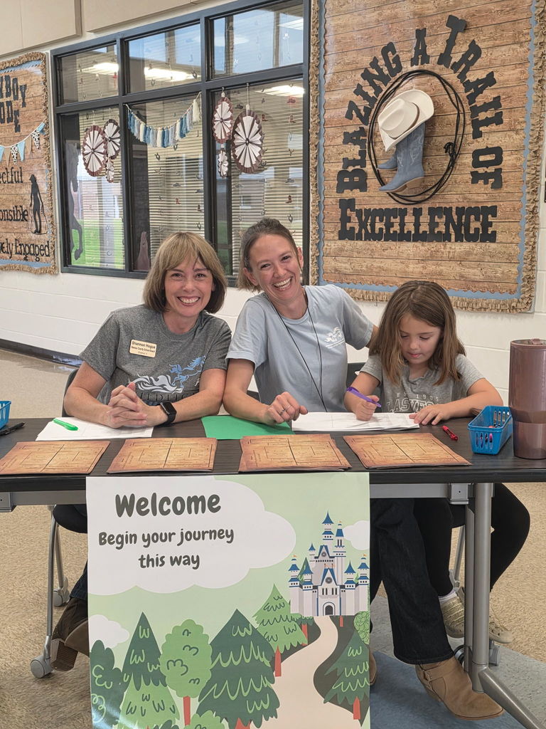 Two staff members and a young student smiling behind a welcome table at Wildlight Elementary, featuring a 'Begin your journey this way' sign for Storybook STEAM Night.