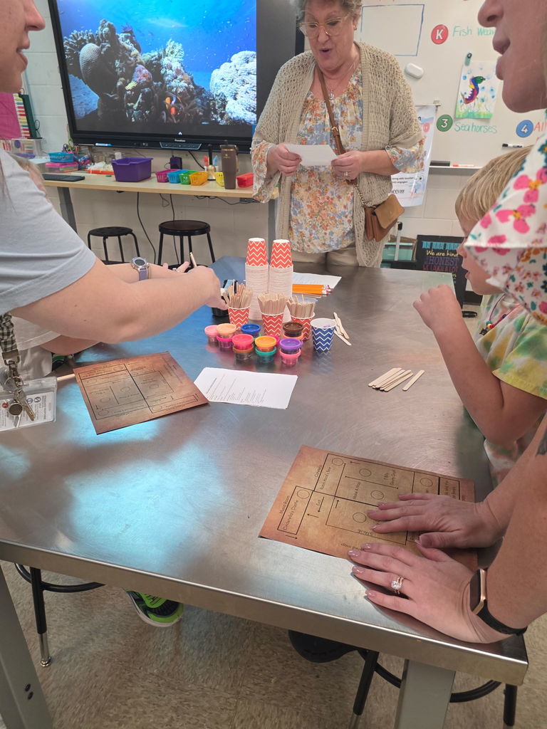 A teacher at Wildlight Elementary assists students at a Storybook STEAM Night station featuring small colorful containers, markers, and measuring tools on a metal table.