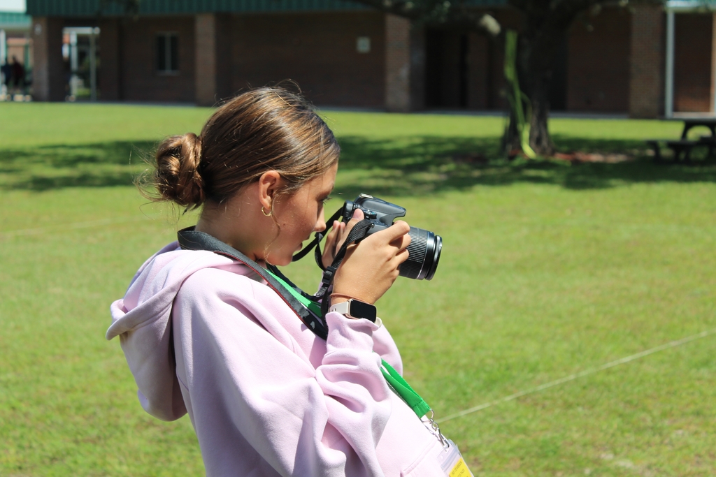 Student journalist looks through the lense of her camera to take a photo. Green grass behind her. 