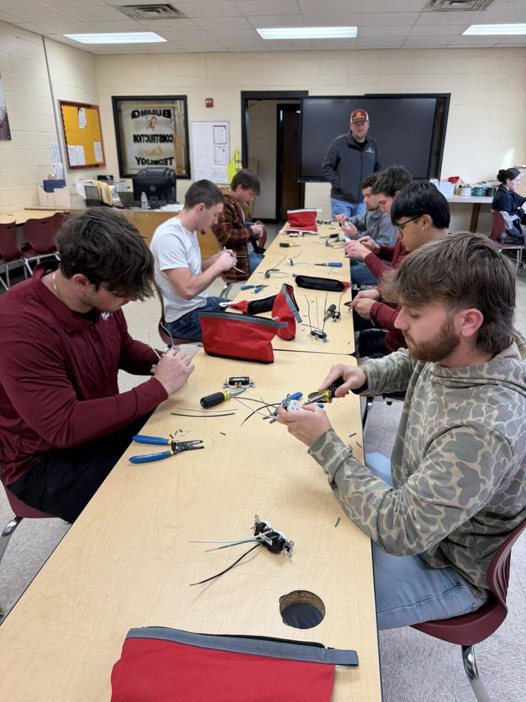 Students in an electricity class at West Nassau High School sit at a long table using wire strippers and tools to practice wiring electrical outlets while their instructor supervises in the classroom.