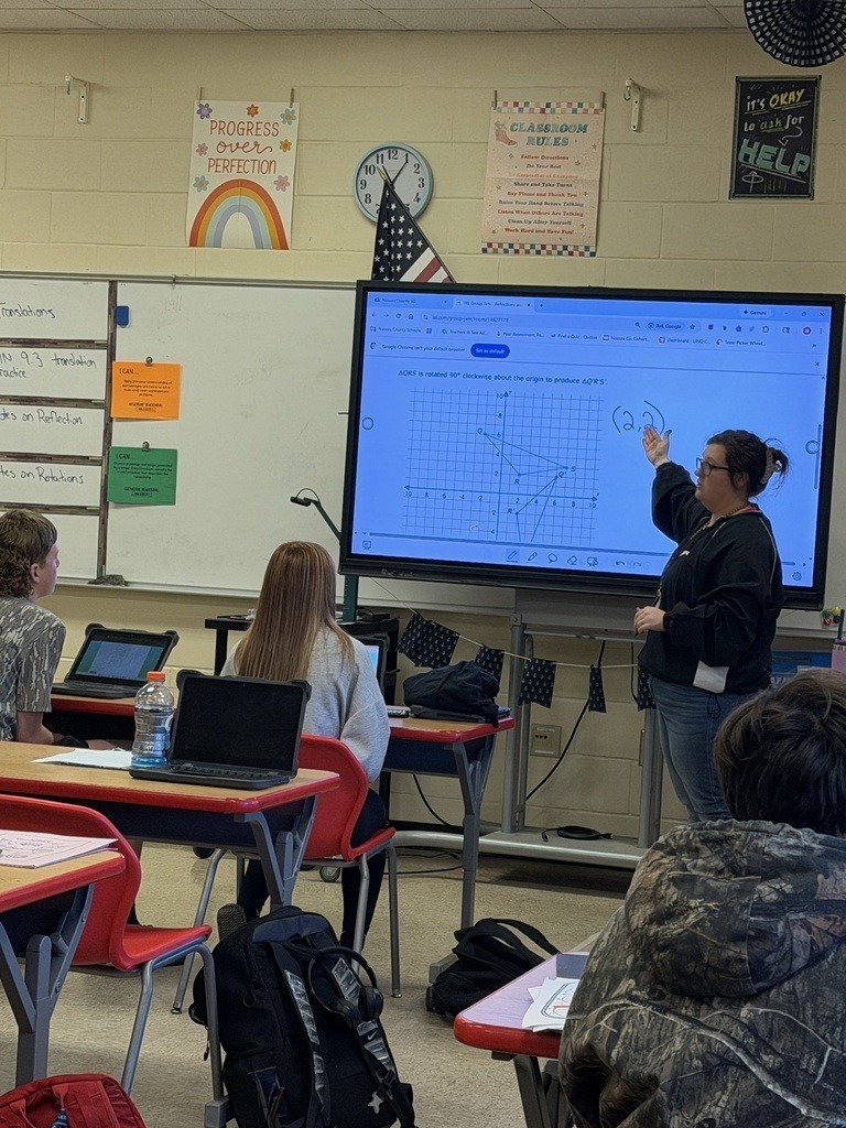A teacher points to a coordinate graph on an interactive classroom display while explaining geometric transformations to students seated at desks with laptops.