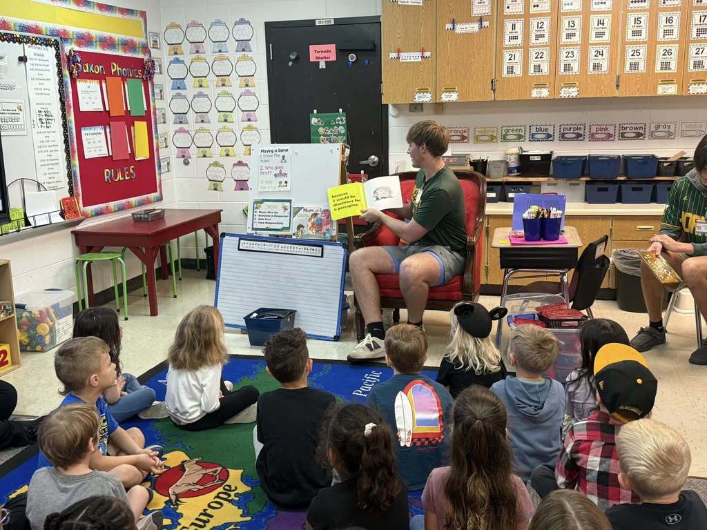 A Yulee High School student reads a picture book to a classroom of primary students sitting on a rug and listening attentively during a guest reading activity.