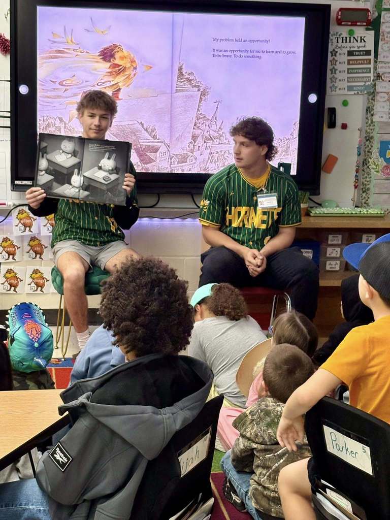 Two Yulee High School students sit at the front of a classroom reading a picture book aloud to a group of young students seated on the floor at Yulee Primary School.