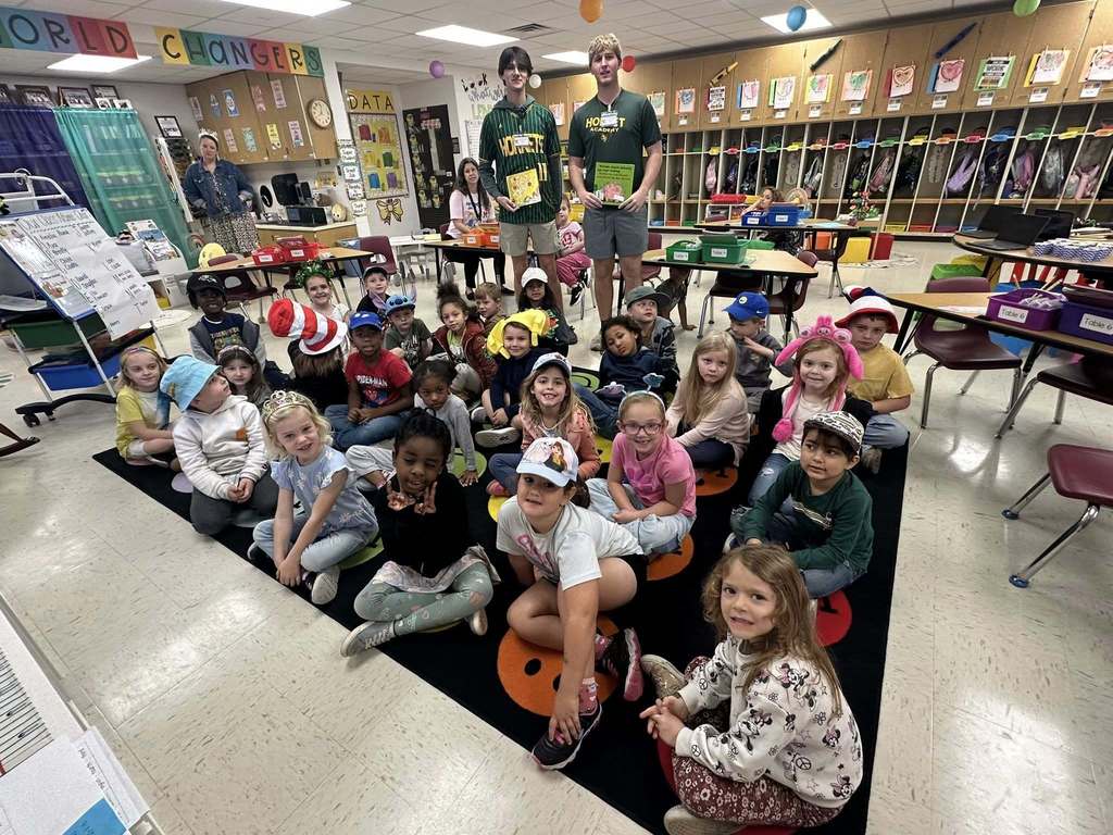 Primary students sit together on a classroom rug while two Yulee High School students stand behind them holding books during a guest reader visit.