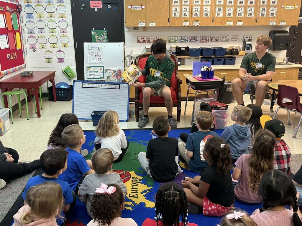 A Yulee High School student reads a children’s book to a classroom of primary students gathered on a rug in front of the reading area.