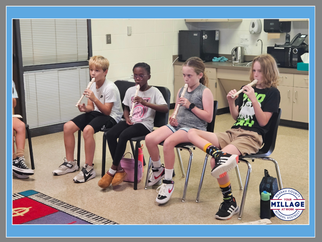 Four elementary students sitting in a row on chairs, each holding and playing a white recorder. They are focused on their music in a classroom with a "Millage at Work" logo in the bottom corner.