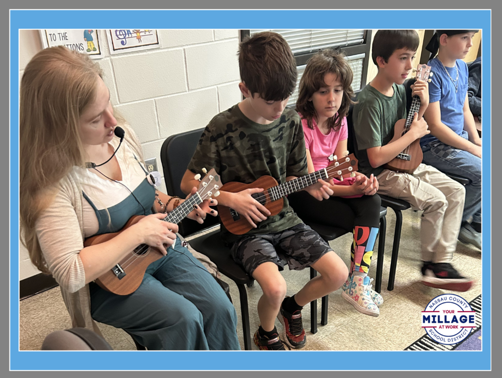 A music teacher and three elementary students sitting together, each holding and playing a ukulele. They are focused on their instruments in a classroom setting with a "Millage at Work" logo visible in the corner.