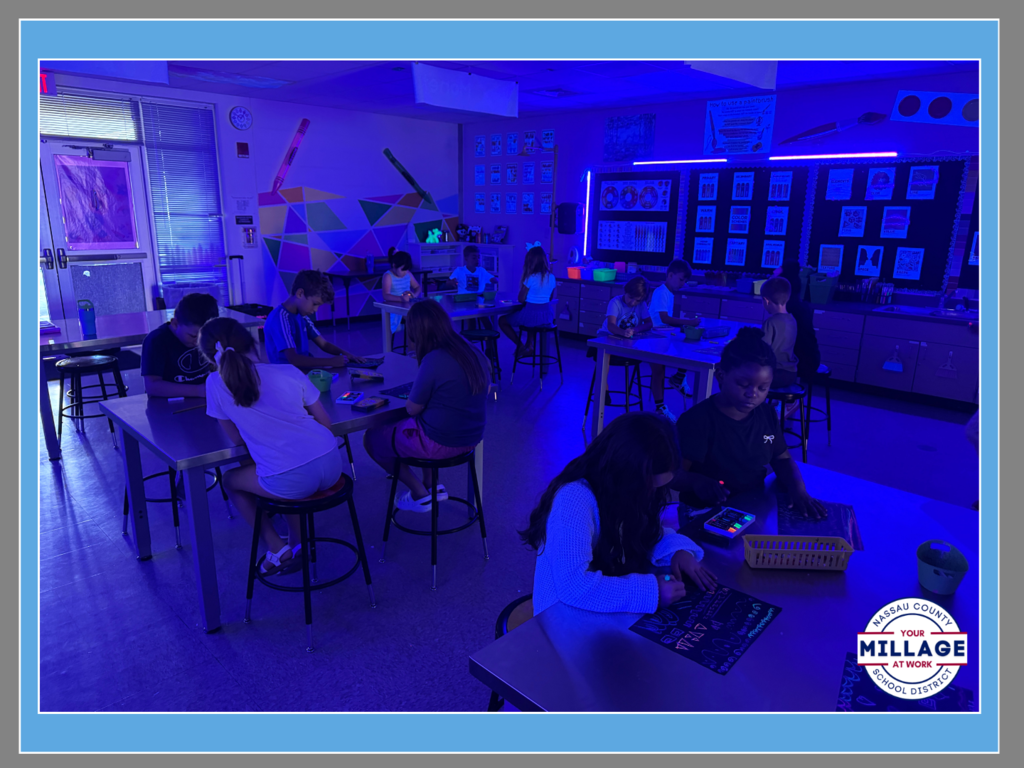 Students working at desks in a classroom illuminated with vibrant blue overhead lighting. The room features a "Millage at Work" logo in the corner and various educational posters on the walls.