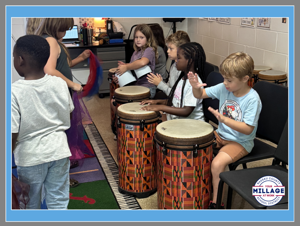 Several elementary students sitting in a music classroom, actively playing large colorful floor drums. A student in the foreground is raised as if mid-beat, while others focus on their instruments.