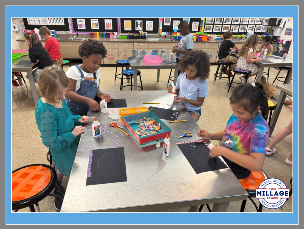 Four elementary students sitting at a classroom table working on an art project. They are using glue and small colorful craft pieces on black paper. A "Millage at Work" logo is in the bottom right corner.