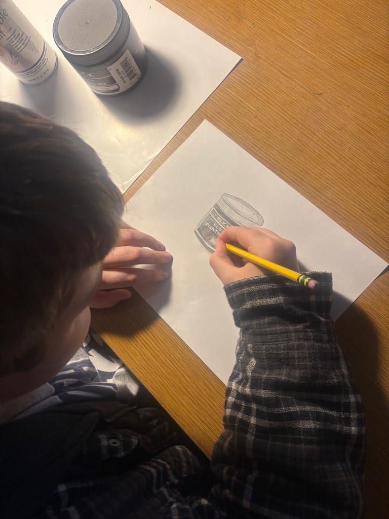 Picture taken from above of a student drawing a can on white paper with a pencil. 