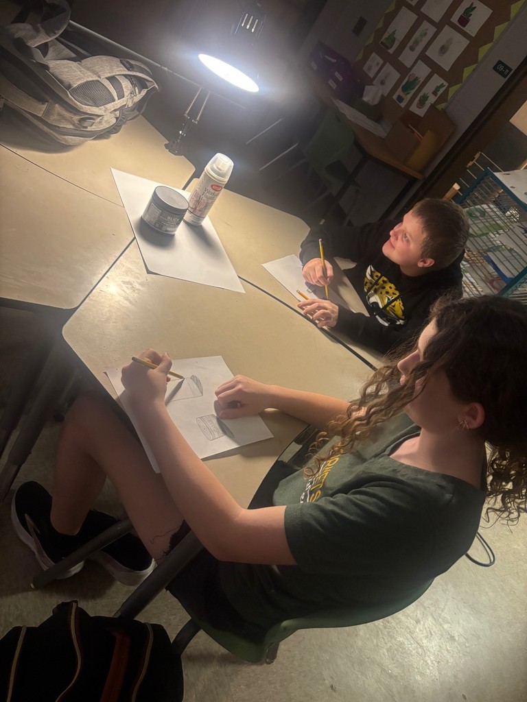 Students draw pictures on white paper as their desks are pushed together under lamp light