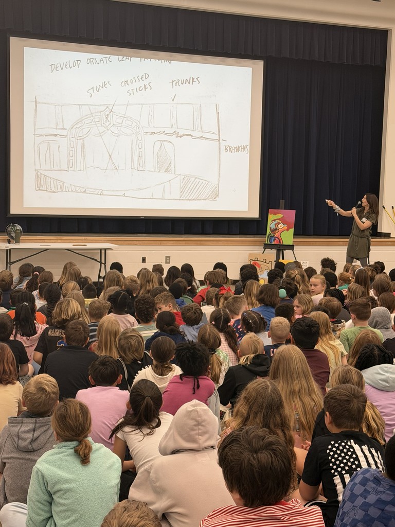 A large group of Wildlight Elementary students sitting on the floor of the school gymnasium, attentively watching an author presentation on a stage with a large projector screen.