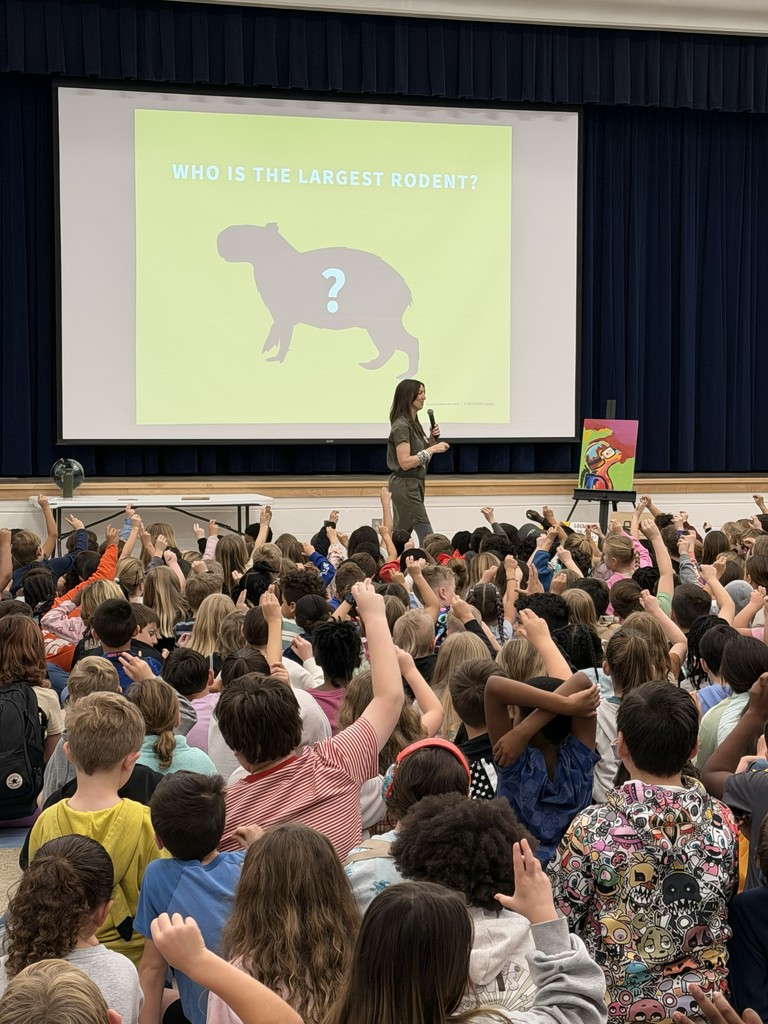 A large group of Wildlight Elementary students sitting on the floor of the school gymnasium, attentively watching an author presentation on a stage with a large projector screen.