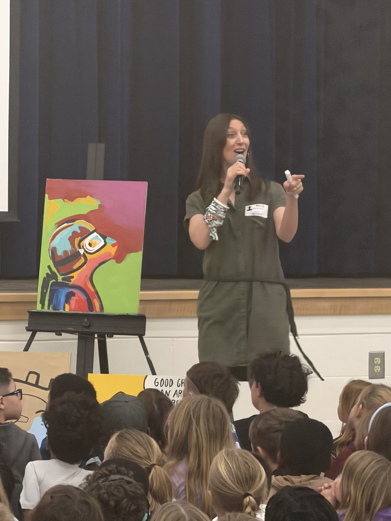 A large group of Wildlight Elementary students sitting on the floor of the school gymnasium, attentively watching an author presentation on a stage with a large projector screen.