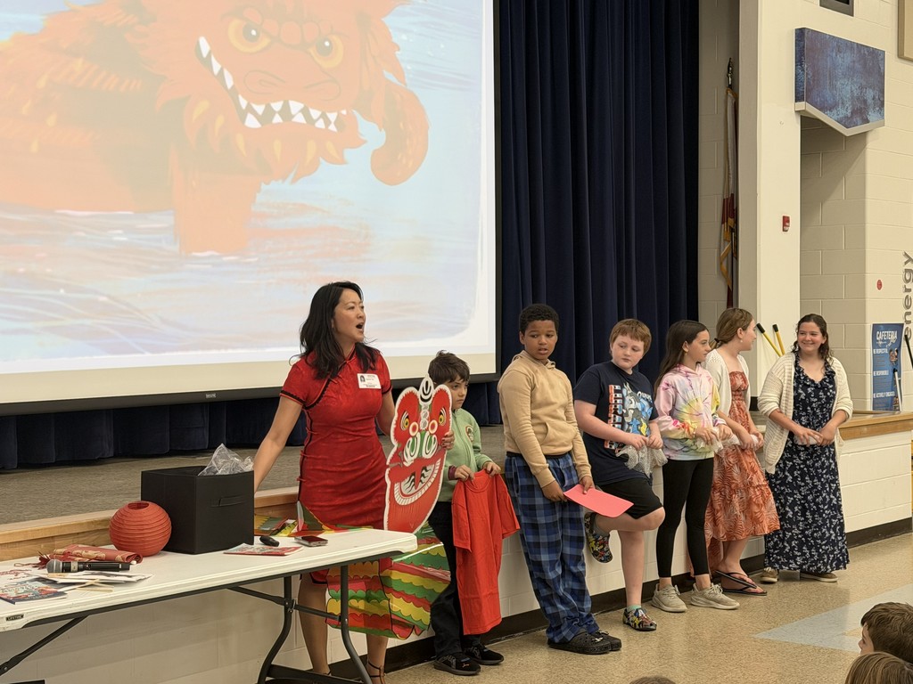 A large group of Wildlight Elementary students sitting on the floor of the school gymnasium, attentively watching an author presentation on a stage with a large projector screen.