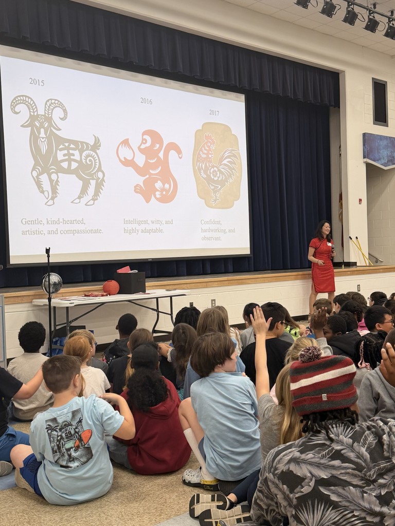 A large group of Wildlight Elementary students sitting on the floor of the school gymnasium, attentively watching an author presentation on a stage with a large projector screen.