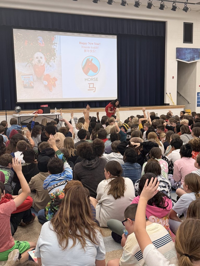 A large group of Wildlight Elementary students sitting on the floor of the school gymnasium, attentively watching an author presentation on a stage with a large projector screen.