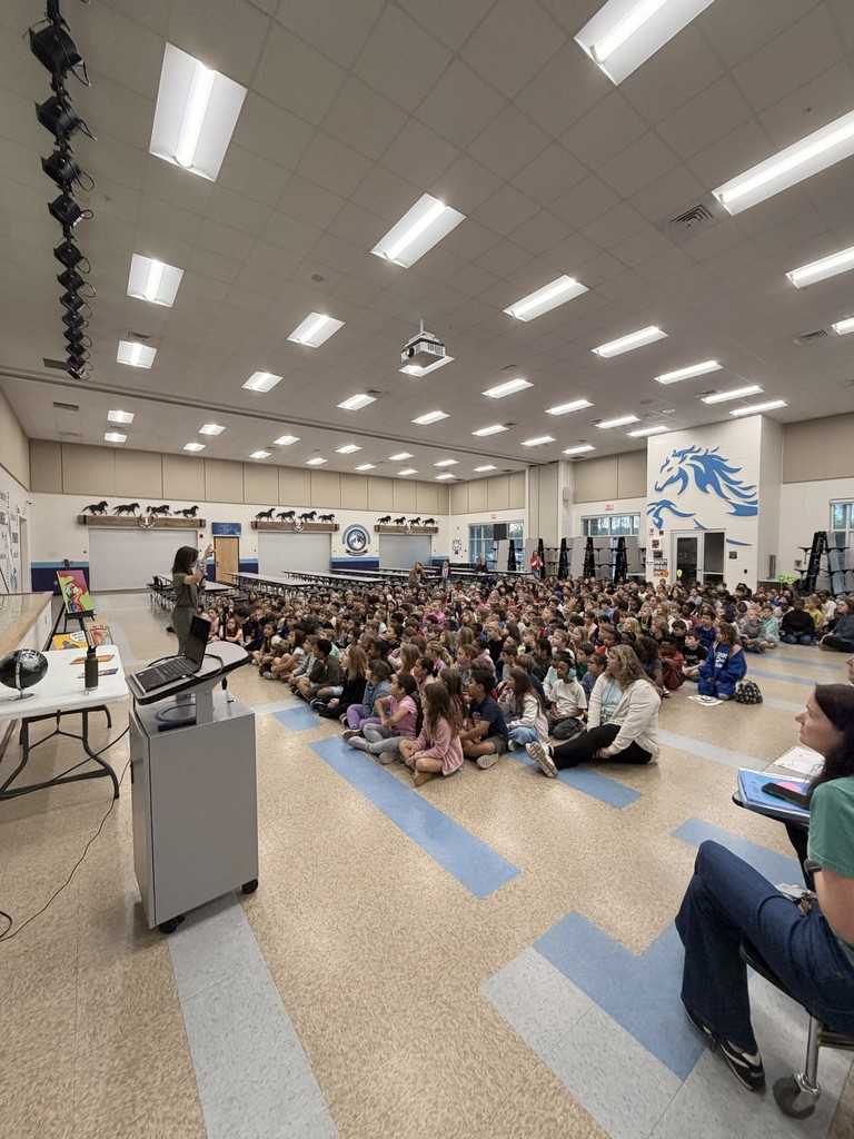 A large group of Wildlight Elementary students sitting on the floor of the school gymnasium, attentively watching an author presentation on a stage with a large projector screen.
