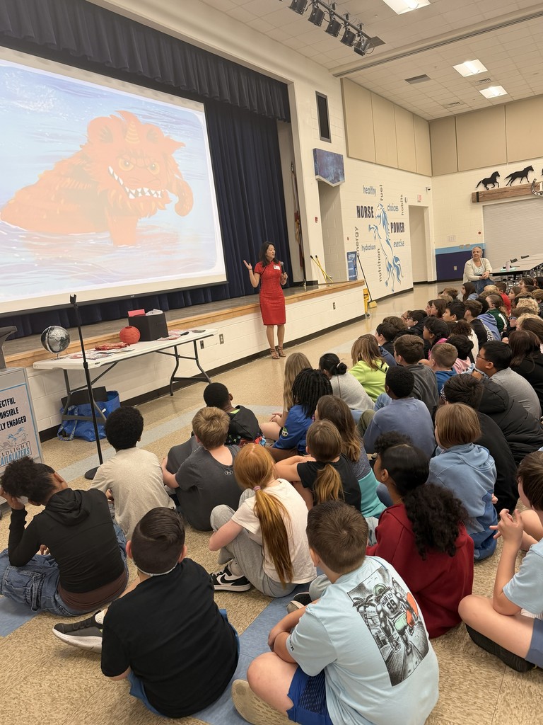 A large group of Wildlight Elementary students sitting on the floor of the school gymnasium, attentively watching an author presentation on a stage with a large projector screen.