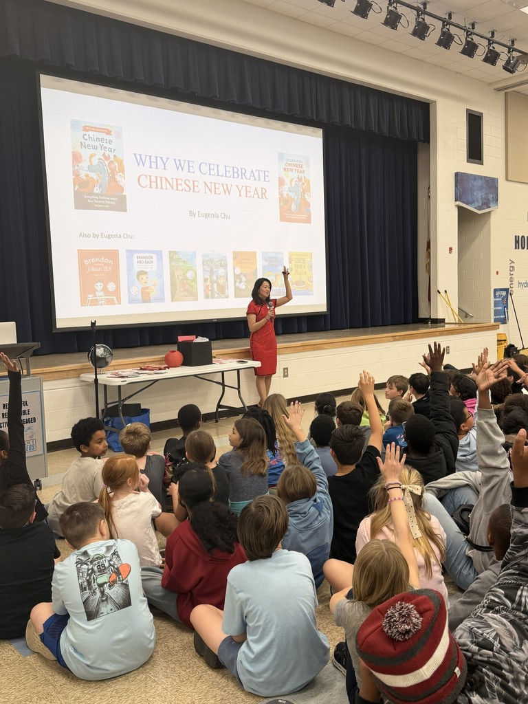 A large group of Wildlight Elementary students sitting on the floor of the school gymnasium, attentively watching an author presentation on a stage with a large projector screen.