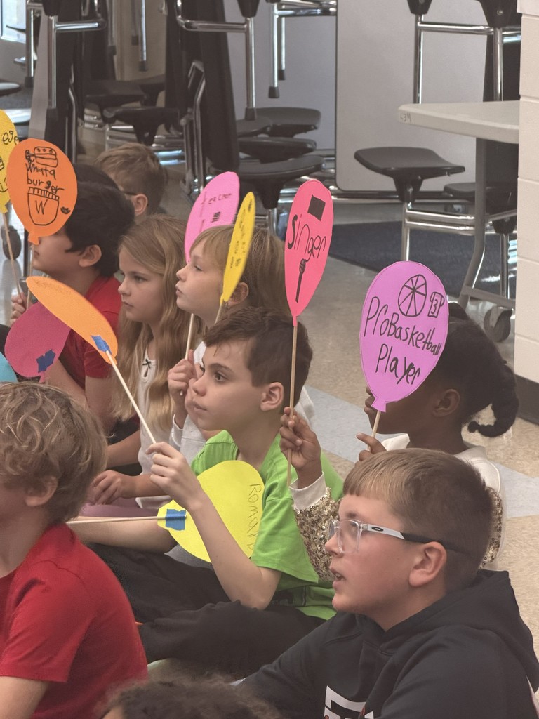 A large group of Wildlight Elementary students sitting on the floor of the school gymnasium, attentively watching an author presentation on a stage with a large projector screen.