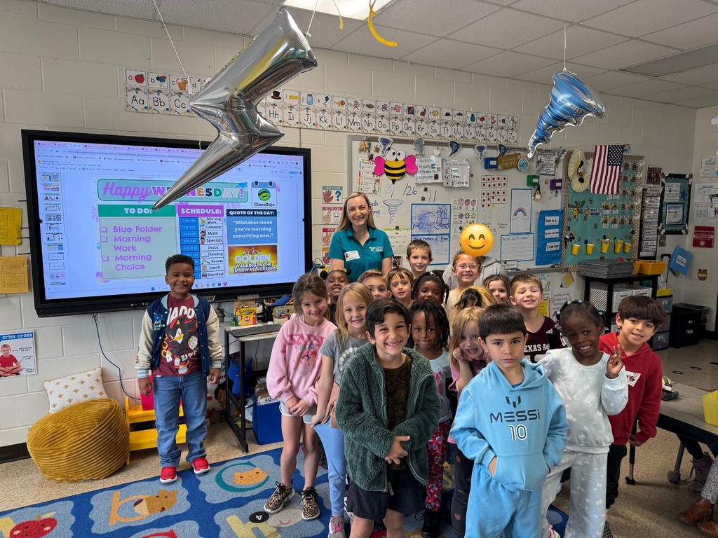 "A group of kindergarten students and their teachers posing for a photo in a classroom with a guest meteorologist. A large screen in the background shows a weather presentation, and the students are gathered on a colorful rug."