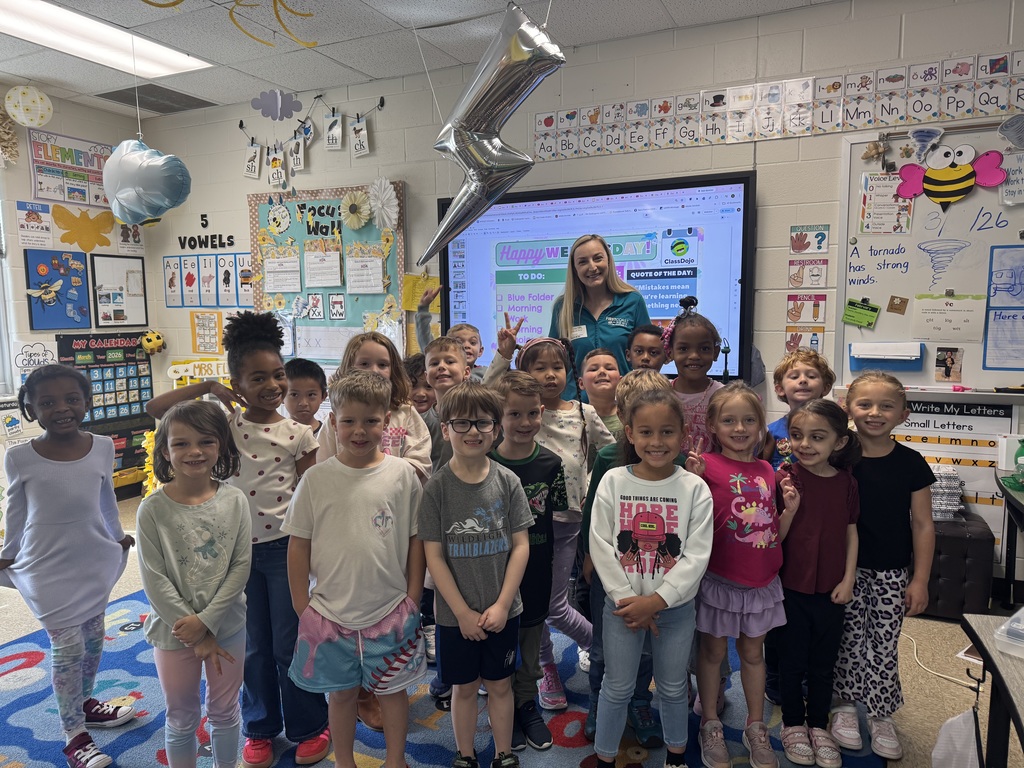 "A group of kindergarten students and their teachers posing for a photo in a classroom with a guest meteorologist. A large screen in the background shows a weather presentation, and the students are gathered on a colorful rug."