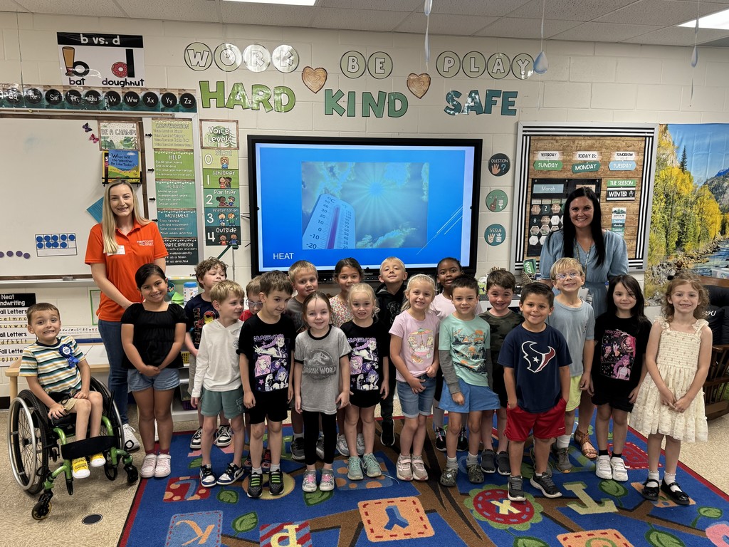 "A group of kindergarten students and their teachers posing for a photo in a classroom with a guest meteorologist. A large screen in the background shows a weather presentation, and the students are gathered on a colorful rug."