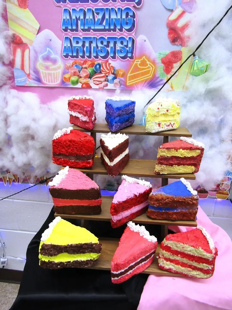 A large collection of colorful, student-made paper-mâché cake slices displayed on a tiered wooden stand. The designs include various 'flavors' like chocolate, rainbow, and strawberry, showcasing different shapes and artistic textures