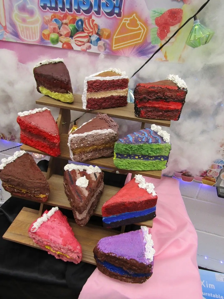 A large collection of colorful, student-made paper-mâché cake slices displayed on a tiered wooden stand. The designs include various 'flavors' like chocolate, rainbow, and strawberry, showcasing different shapes and artistic textures