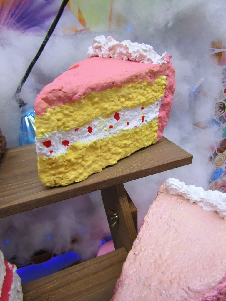 A detailed close-up of a handmade 3D cake slice art project. The 'cake' features a yellow base, a white middle layer with red 'sprinkles,' and pink frosting on top, all textured with a paper-mâché and paint finish