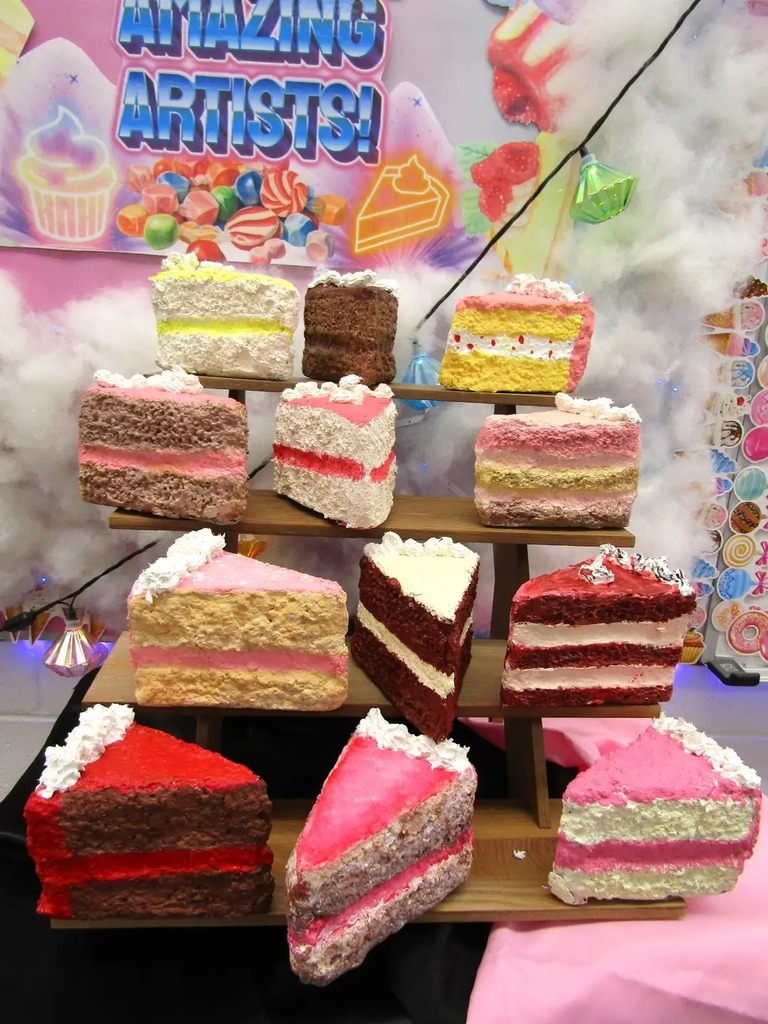 A display of colorful, handmade 3D art projects shaped like slices of cake. The 'cakes' feature various designs, including pink frosting, chocolate layers, and red velvet, all created using paper-mâché and fabric by 5th-grade students