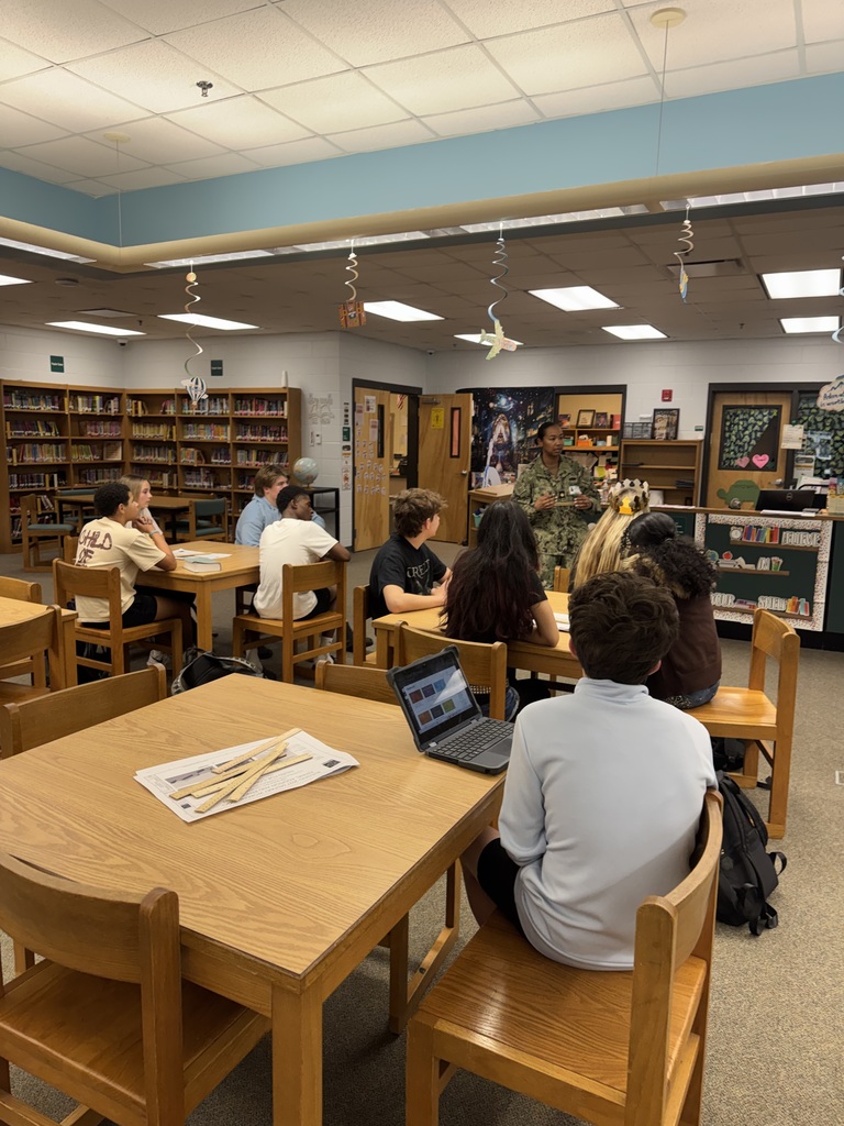 Students sit in a library seated at brown tables while listening to a speaker in military uniform.
