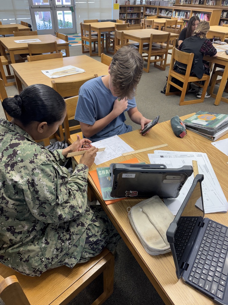 Mrs. King works with students in her military uniform at their table in the library. Laptops, rulers and worksheets cover the table. 