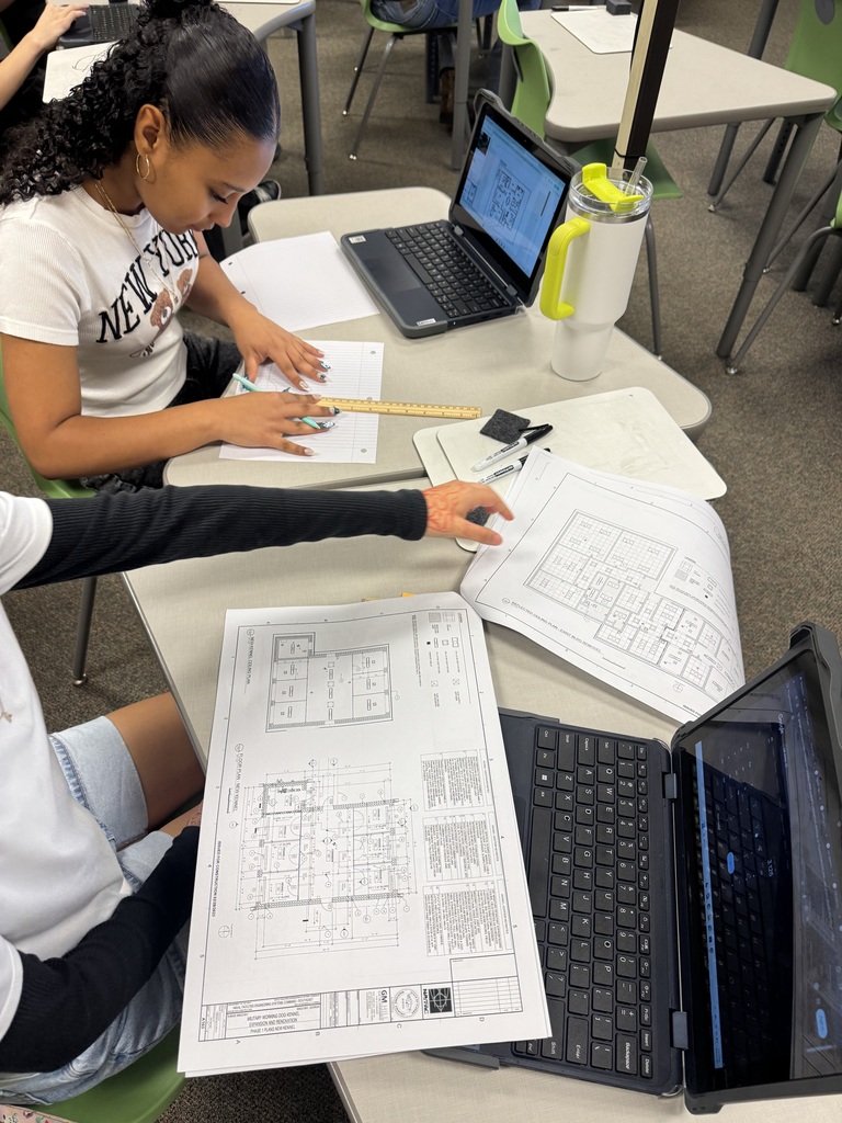 Two students work on blueprints while sitting at their white topped desks.