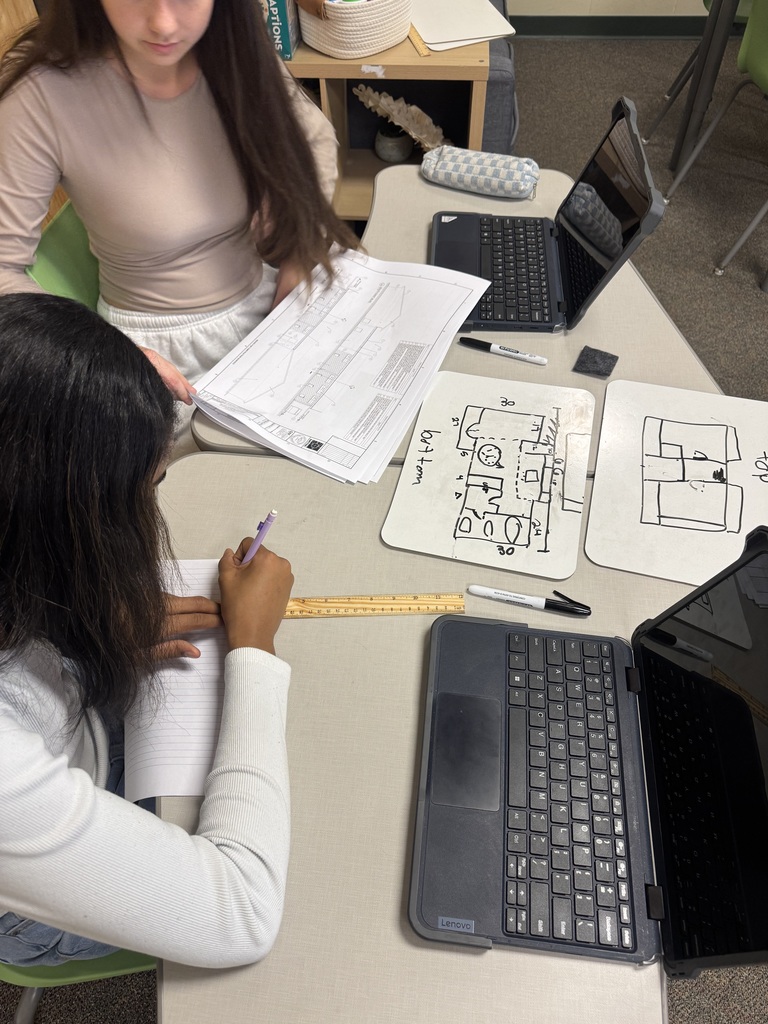 Two students work at a table with a ruler and laptops designing blueprints.