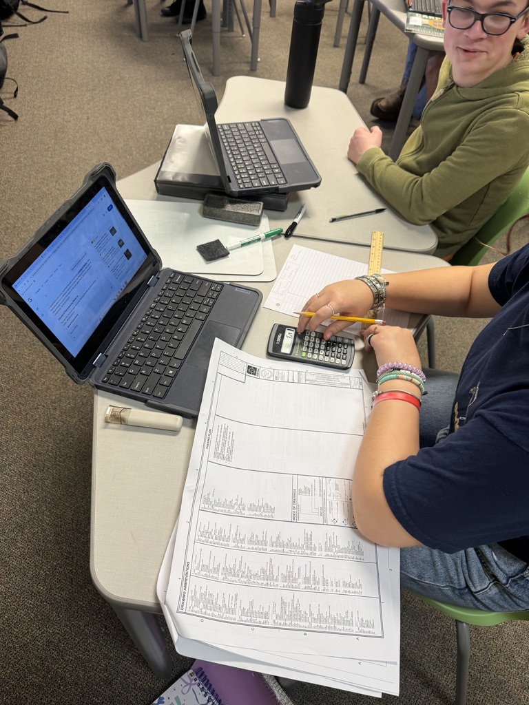 Two students work at a table with a ruler and laptops designing blueprints.