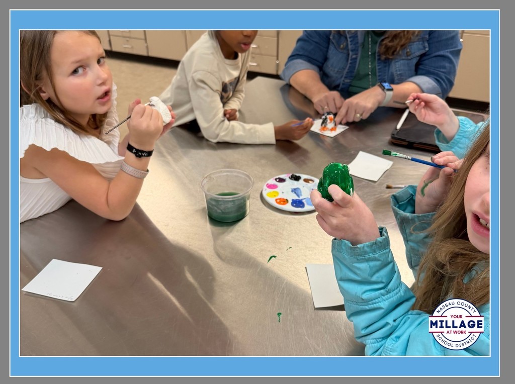 There young students at an art table focus on painting small projects. One student in a blue jacket holds a paintbrush over a green object, while a circular paint palette with various colors sits in the center of the table.