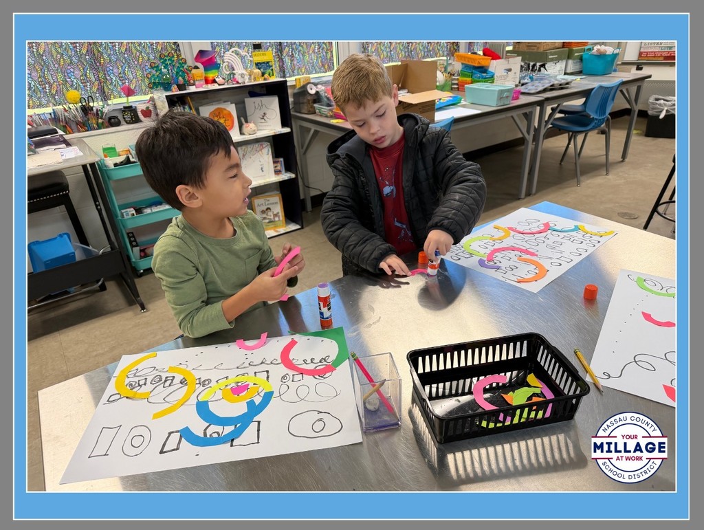 Two young students sit at a classroom table working on art projects. They are using paper to make colorful, swirling abstract patterns on white paper. A black basket of supplies and a small "Millage" logo are visible on the table.
