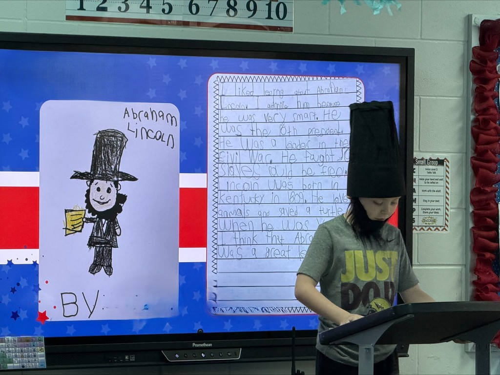 A young student stands at a classroom podium, presenting a research project. In the background, a large digital screen displays a handwritten essay about a U.S. President next to an image of the president..
