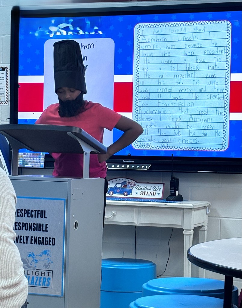 A young student stands at a classroom podium, presenting a research project. In the background, a large digital screen displays a handwritten essay about a U.S. President next to an image of the president..