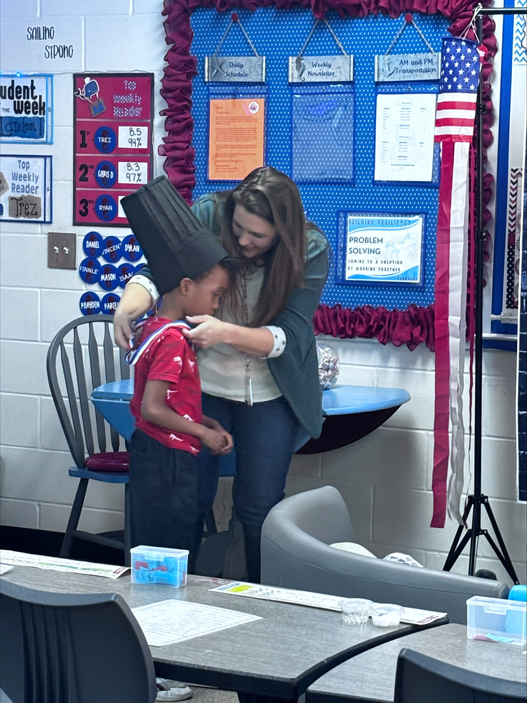 A young student stands at a classroom podium, presenting a research project. In the background, a large digital screen displays a handwritten essay about a U.S. President next to an image of the president..