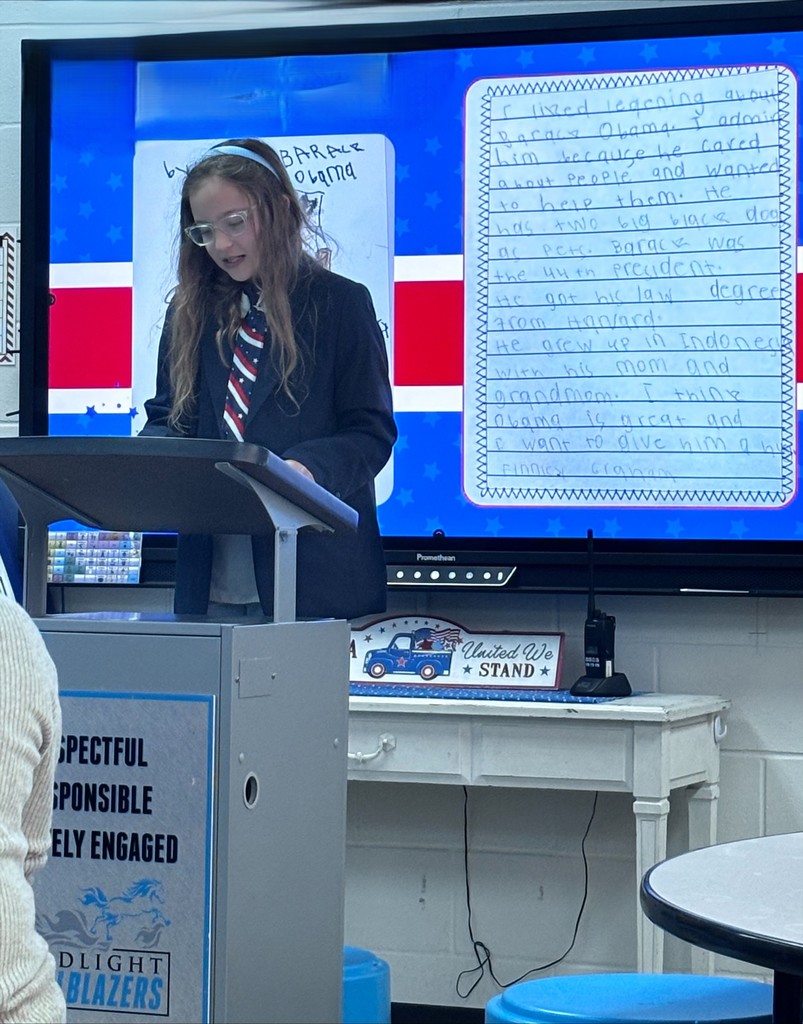 A young student stands at a classroom podium, presenting a research project. In the background, a large digital screen displays a handwritten essay about a U.S. President next to an image of the president..