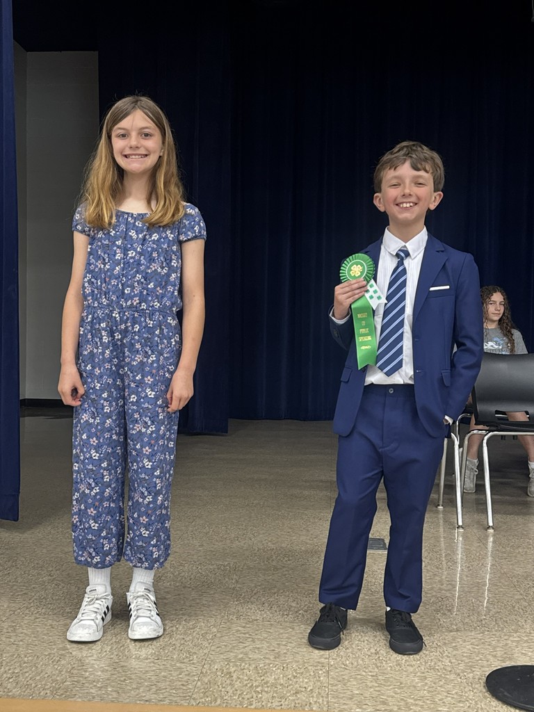 Two elementary students stand on a school stage in front of a blue curtain. A student in a blue and white floral jumpsuit stands next to a student in a navy blue suit who is smiling and holding up a green award ribbon.