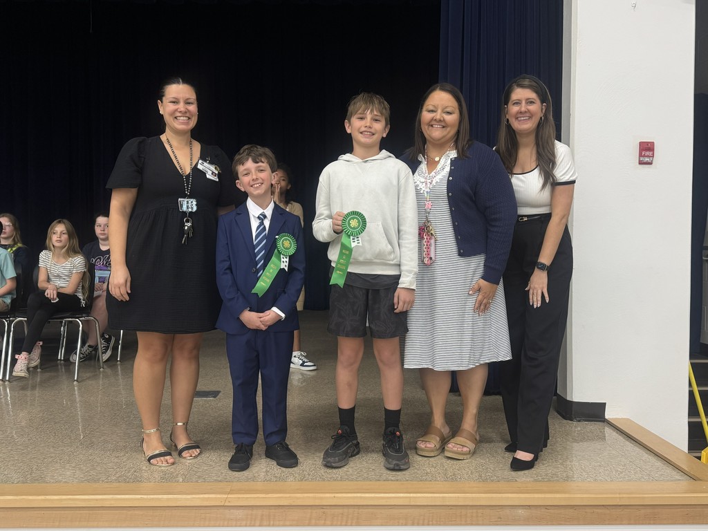 Two students stand between three teachers on a school stage. The students are wearing semi-formal attire and holding green award ribbons. Everyone is smiling for the camera in front of a dark blue stage curtain.