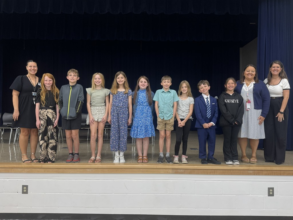 A large group of nine elementary students and three teachers posing together on a school stage. The students are dressed in a variety of nice outfits, including dresses and suits, and are lined up in front of a dark blue curtain for a group photo.