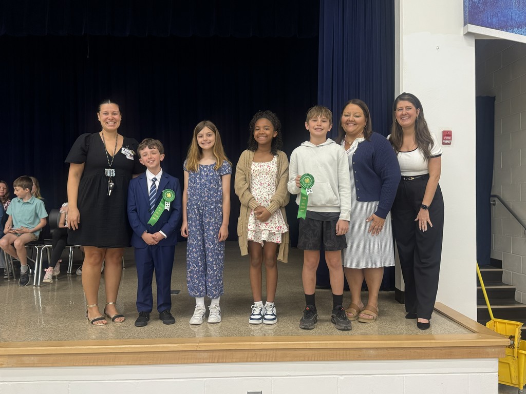 A group photo of five elementary students standing with three teachers on a school stage. The students are dressed in semi-formal clothing, and two of the students are holding green award ribbons. Everyone is smiling in front of the dark blue stage curtains.