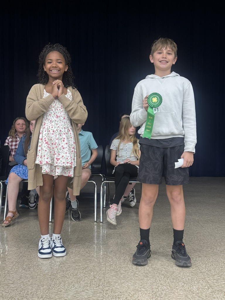 An elementary student inTwo students stand on a school stage, smiling and proudly holding a green award ribbon. Other students and a set of chairs are visible in the blurred background.